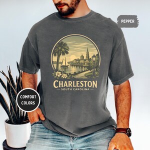 May include: Dark gray t-shirt featuring a circular graphic of Charleston, South Carolina, with a palm tree, sailboat, and city skyline. The text "Charleston South Carolina" is printed below the image. The shirt has short sleeves and a crew neck.