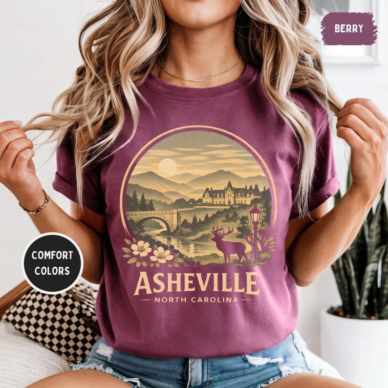 May include: Berry-colored t-shirt featuring a scenic illustration of Asheville, North Carolina. The design includes a bridge, mountains, a deer, and the text "Asheville - North Carolina". The shirt also has the words "Comfort Colors" printed on it.