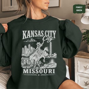 May include: Forest green sweatshirt featuring a white graphic of a rodeo rider on a bucking bronco, with the words "KANSAS CITY" above "City" and "MISSOURI" below. The design includes city skyline and architectural elements.