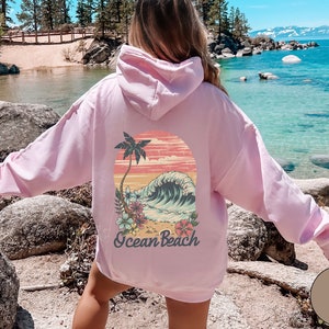 Op de afbeelding: Een lichtroze hoodie met een capuchon. De achterkant van de hoodie heeft een kleurrijke afbeelding van een palmboom, oceaangolven en de tekst "Ocean Beach". Het ontwerp is geplaatst tegen een zonsondergangachtergrond.