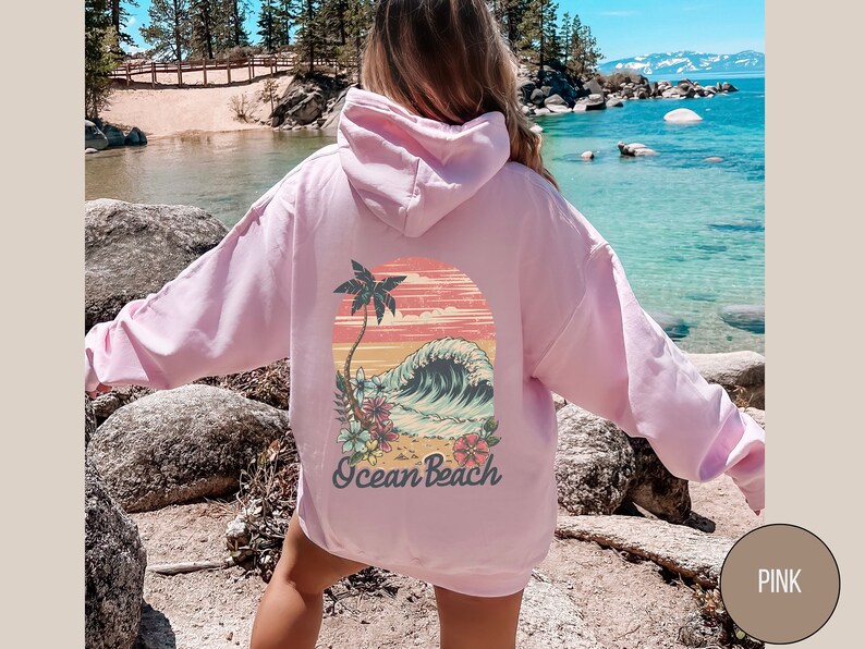 Op de afbeelding: Een lichtroze hoodie met een capuchon. De achterkant van de hoodie heeft een kleurrijke afbeelding van een palmboom, oceaangolven en de tekst "Ocean Beach". Het ontwerp is geplaatst tegen een zonsondergangachtergrond.