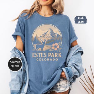 May include: Blue jean-colored t-shirt with a circular graphic featuring a mountain range, deer, and flower, with the text "Estes Park Colorado." The shirt has rolled-up sleeves and is part of the "Comfort Colors" collection.