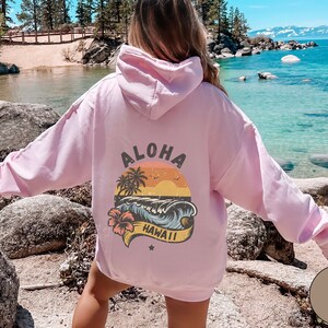 Puede incluir: Sudadera rosa con un estampado gr&aacute;fico de una puesta de sol sobre una playa con palmeras y las palabras "Aloha Hawaii".