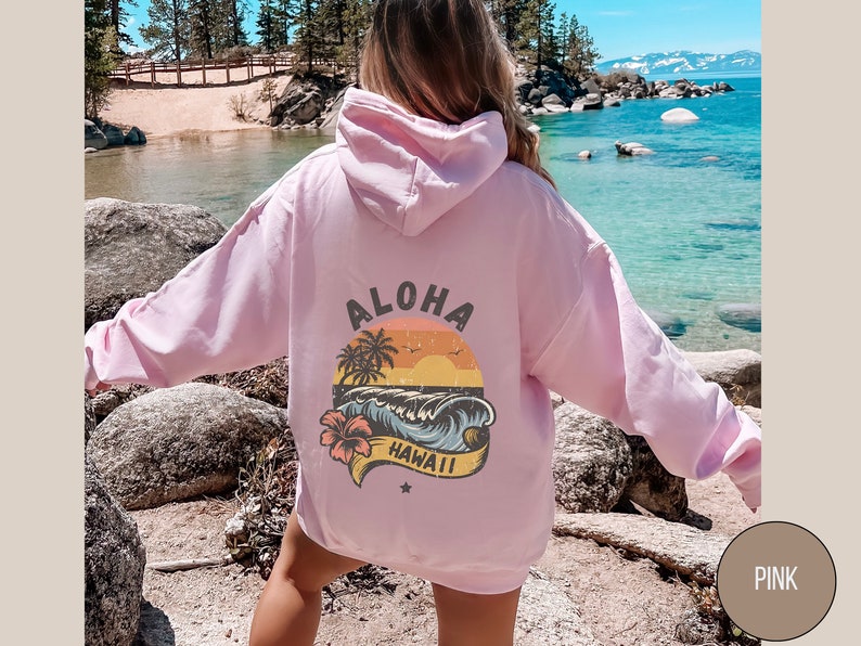 Puede incluir: Sudadera rosa con un estampado gr&aacute;fico de una puesta de sol sobre una playa con palmeras y las palabras "Aloha Hawaii".