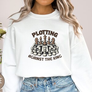 May include: White sweatshirt with a brown graphic of a chessboard with the text "Plotting Against The King" on it.