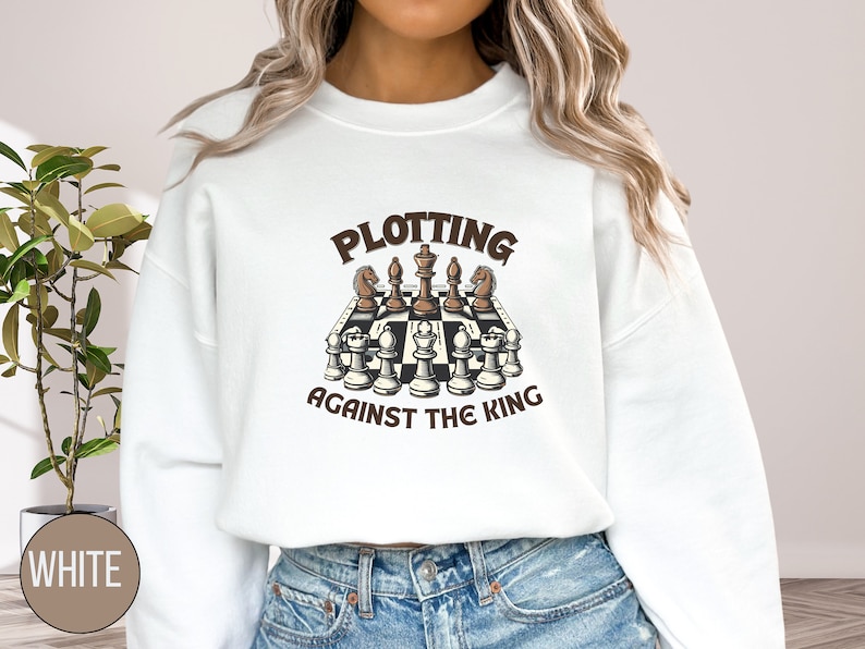 May include: White sweatshirt with a brown graphic of a chessboard with the text "Plotting Against The King" on it.