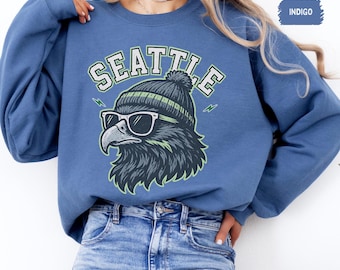 Könnte beinhalten: Indigoblaues Sweatshirt mit einer Grafik eines stilisierten Adlers mit Sonnenbrille und Mütze. Das Wort "SEATTLE" ist in Grün und Weiß über dem Adler aufgedruckt. Das Sweatshirt hat lange Ärmel und einen Rundhalsausschnitt.