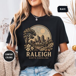 May include: Black t-shirt with a circular graphic featuring a cityscape, tree, and floral elements. The text "RALEIGH NORTH CAROLINA" is displayed below the graphic. The shirt also has the text "COMFORT COLORS" and "BLACK".