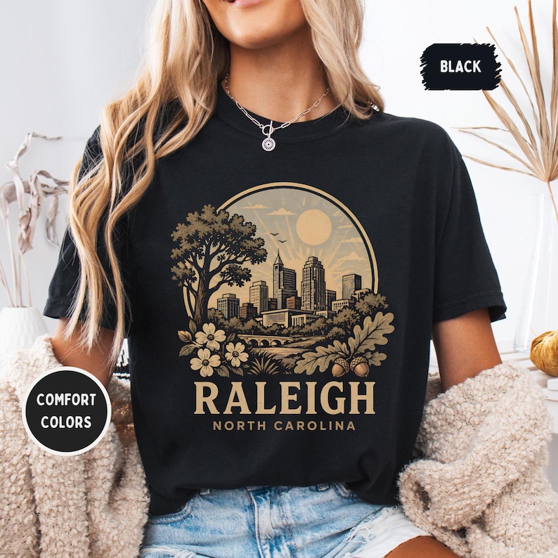 May include: Black t-shirt with a circular graphic featuring a cityscape, tree, and floral elements. The text "RALEIGH NORTH CAROLINA" is displayed below the graphic. The shirt also has the text "COMFORT COLORS" and "BLACK".