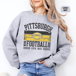 May include: Sport grey sweatshirt with a distressed graphic that reads "PITTSBURGH FOOTBALL ESTD. 1933". The design features a football and black and yellow stripes, representing the team colors.