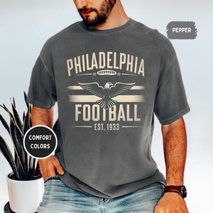 May include: A dark gray t-shirt with a vintage-style graphic design featuring the text "Philadelphia Football Est. 1933" and an eagle with outstretched wings.