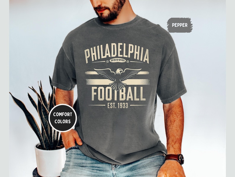 May include: A dark gray t-shirt with a vintage-style graphic design featuring the text "Philadelphia Football Est. 1933" and an eagle with outstretched wings.