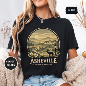 May include: Black t-shirt with a circular graphic featuring a mountain landscape, bridge, and deer. The text "Asheville - North Carolina" is below the graphic. The shirt also has the text "Comfort Colors" and "Black".