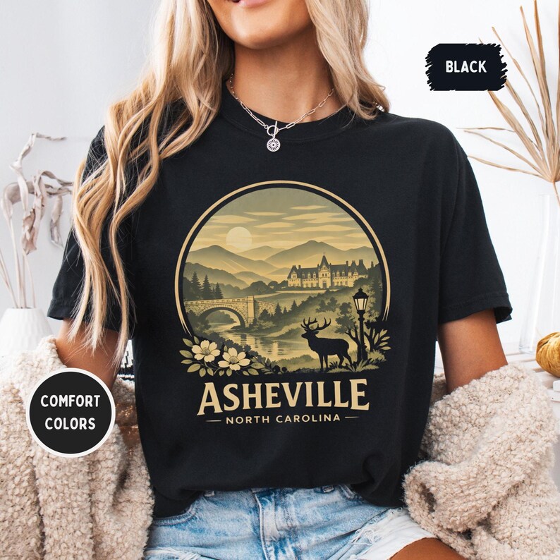 May include: Black t-shirt with a circular graphic featuring a mountain landscape, bridge, and deer. The text "Asheville - North Carolina" is below the graphic. The shirt also has the text "Comfort Colors" and "Black".