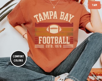Puede incluir: Una camiseta de color óxido con las palabras "TAMPA BAY FOOTBALL" y "ESTD. 1976" impresas en un estilo vintage. El diseño incluye un gráfico de fútbol americano y rayas horizontales en tonos rojos, naranjas y amarillos. La camiseta es de la colección Comfort Colors.