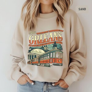 May include: A beige sweatshirt with a graphic print of a vintage-style streetcar in New Orleans. The text "New Orleans" is in a retro font.