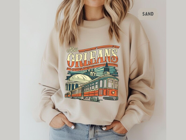 May include: A beige sweatshirt with a graphic print of a vintage-style streetcar in New Orleans. The text "New Orleans" is in a retro font.