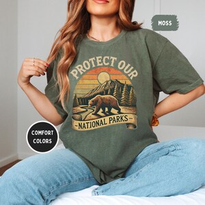 May include: A green t-shirt with a vintage-style graphic of a bear walking through a forest with the text "Protect Our National Parks".
