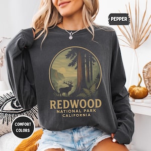 Pode incluir: Camisola cinza escuro com um gráfico circular com um veado, árvores e o texto "Redwood National Park California". A palavra "Pepper" está impressa no canto superior direito. A camisola faz parte da coleção "Comfort Colors".