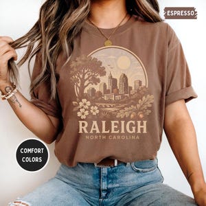 May include: Espresso-colored t-shirt featuring a circular graphic of the Raleigh, North Carolina skyline, with the city name and state printed below. The shirt has rolled-up sleeves and is made of a soft, comfortable material. The words "Comfort Colors" are printed on a circular sticker.