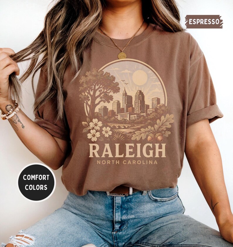 May include: Espresso-colored t-shirt featuring a circular graphic of the Raleigh, North Carolina skyline, with the city name and state printed below. The shirt has rolled-up sleeves and is made of a soft, comfortable material. The words "Comfort Colors" are printed on a circular sticker.