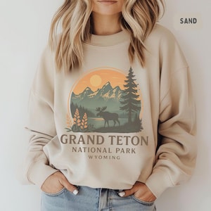 Könnte beinhalten: Sandfarbenes Sweatshirt mit einer Grafik des Grand Teton Nationalparks in Wyoming. Das Design zeigt Berge, einen See, einen Elch und den Text "GRAND TETON NATIONAL PARK WYOMING" in einem Kreis.