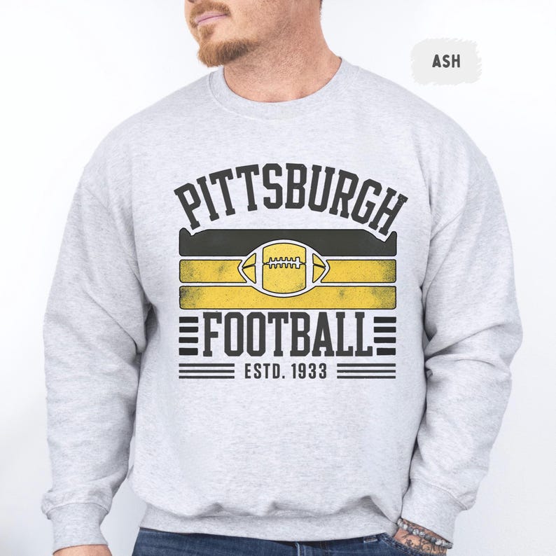 May include: Ash-colored sweatshirt with the text "PITTSBURGH FOOTBALL ESTD. 1933" in black and yellow. The design includes a football graphic and horizontal stripes. The sweatshirt is a crew neck style.