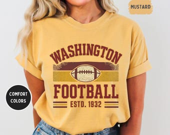 Op de afbeelding: Mosterdkleurig T-shirt met de woorden "WASHINGTON FOOTBALL" en een voetbalafbeelding. Het shirt bevat ook "ESTD. 1932". Het shirt heeft een comfortabele kleur en wordt gedragen met een blauwe jeans.