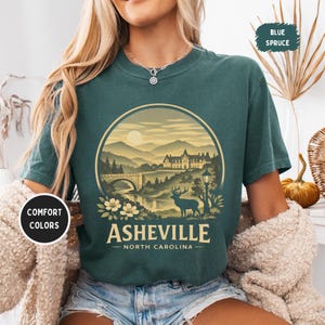 May include: A teal-colored t-shirt featuring a circular graphic of Asheville, North Carolina. The design includes a bridge, mountains, a deer, and the text "Asheville - North Carolina". The shirt also has the text "Comfort Colors" and "Blue Spruce".