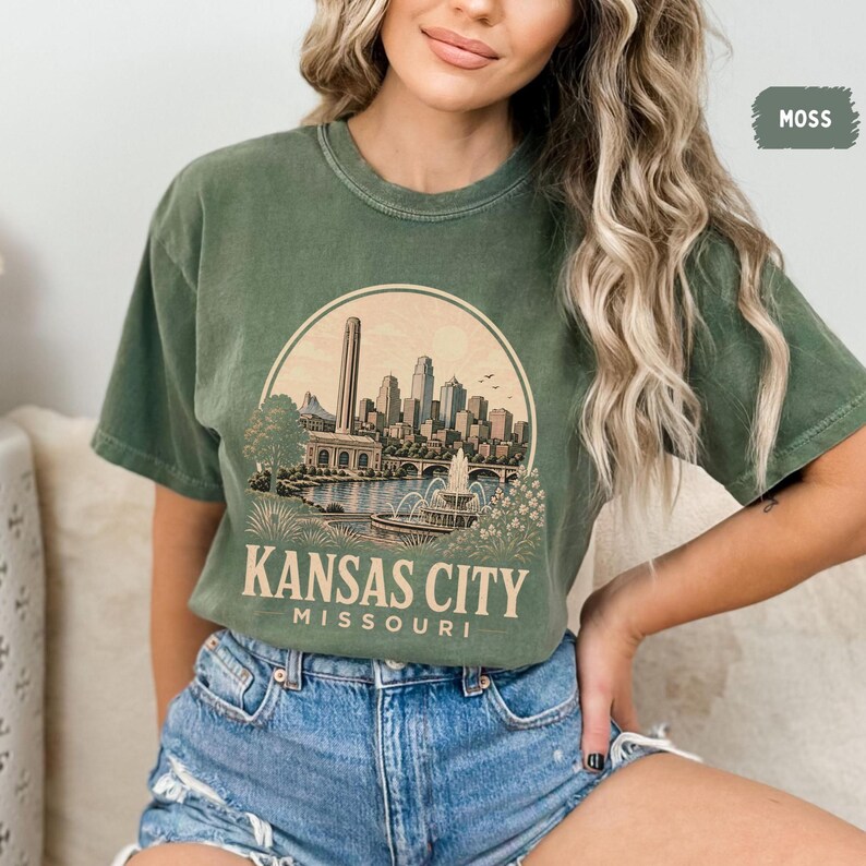 May include: Moss green t-shirt featuring a circular graphic of the Kansas City skyline, a fountain, and the text "Kansas City Missouri". The shirt has a crew neck and short sleeves.