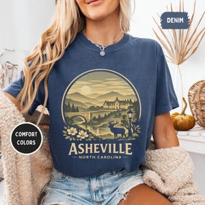 May include: Denim blue t-shirt featuring a circular graphic of Asheville, North Carolina. The design includes a bridge, mountains, a deer, and the text "Asheville - North Carolina". The shirt has the words "Comfort Colors" on a white circle.