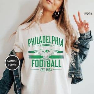May include: White t-shirt with green text that reads "Philadelphia Football Est. 1933". The text is surrounded by a green and white stripe design. A green eagle with outstretched wings is centered above the text.