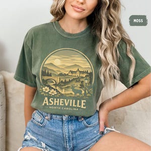 May include: Moss green t-shirt featuring a circular graphic of Asheville, North Carolina. The design includes a bridge, mountains, a deer, and the text "Asheville North Carolina". The shirt has a crew neck and short sleeves.