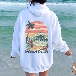 Op de afbeelding: Witte hoodie met een retro afbeelding op de achterkant. De afbeelding toont een zonsondergang, palmboom, oceaangolf en bloemen, met de woorden "Ocean Beach" eronder. De hoodie is effen wit.