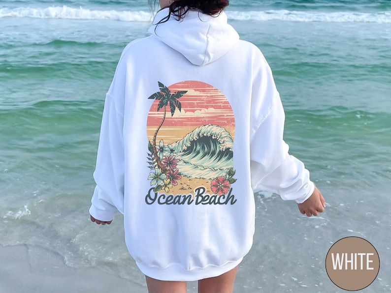 Op de afbeelding: Witte hoodie met een retro afbeelding op de achterkant. De afbeelding toont een zonsondergang, palmboom, oceaangolf en bloemen, met de woorden "Ocean Beach" eronder. De hoodie is effen wit.