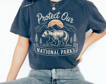 May include: A navy blue t-shirt with a vintage-style graphic of a bear walking through a mountain landscape with a sunset in the background. The text "Protect Our National Parks" is printed below the graphic.
