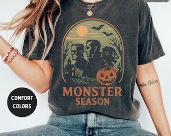May include: Dark grey t-shirt with a Halloween-themed graphic. The design features Frankenstein, Dracula, and a werewolf, with a pumpkin and the words "Monster Season". The shirt has a vintage, worn look.