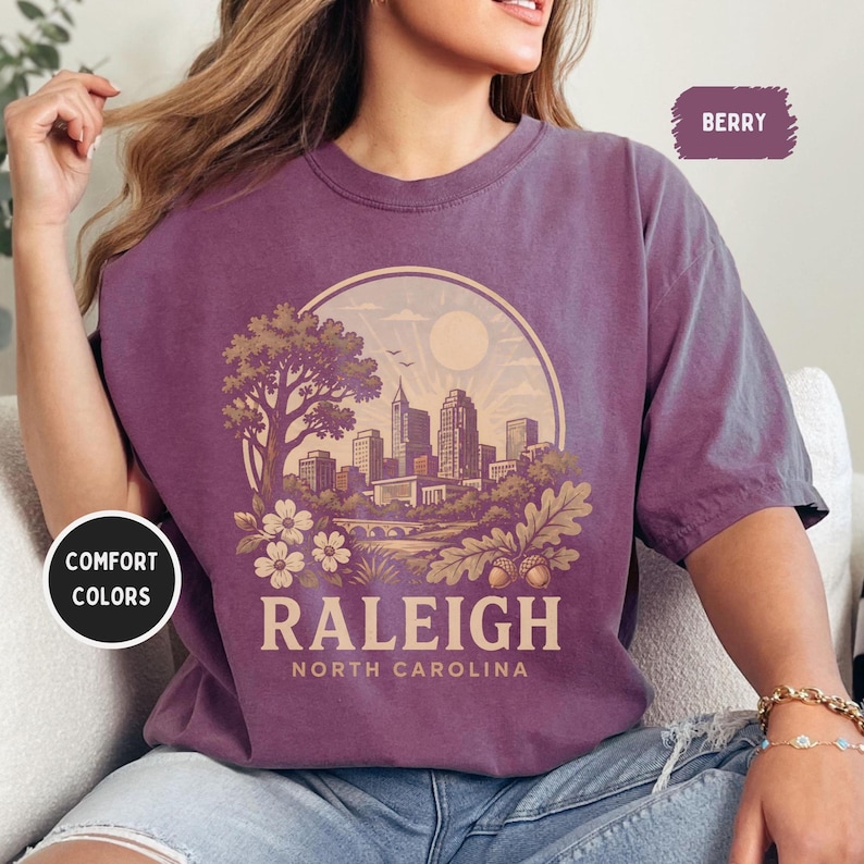 May include: Berry-colored t-shirt featuring a circular graphic of the Raleigh, North Carolina skyline. The design includes a tree, flowers, and the text "RALEIGH NORTH CAROLINA". The shirt has short sleeves and a crew neck. The words "Comfort Colors" are also visible.