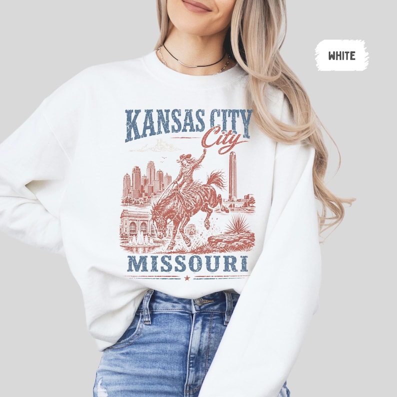 May include: White sweatshirt with a graphic design featuring the text "Kansas City Missouri". The design includes an illustration of a person riding a bucking bronco, with city skyline and landmark details.