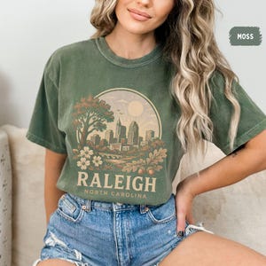 May include: Moss green t-shirt featuring a circular graphic of the Raleigh, North Carolina skyline, with the city name and state printed below. The design includes a tree, flowers, and a bridge. The shirt is made of a soft material.