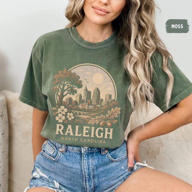 May include: Moss green t-shirt featuring a circular graphic of the Raleigh, North Carolina skyline, with the city name and state printed below. The design includes a tree, flowers, and a bridge. The shirt is made of a soft material.