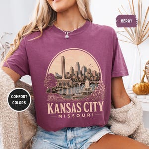May include: Berry-colored t-shirt featuring a circular graphic of the Kansas City skyline, including a tall tower, buildings, a river, and a fountain. The text "Kansas City Missouri" is below the graphic. The shirt has short sleeves and a crew neck.
