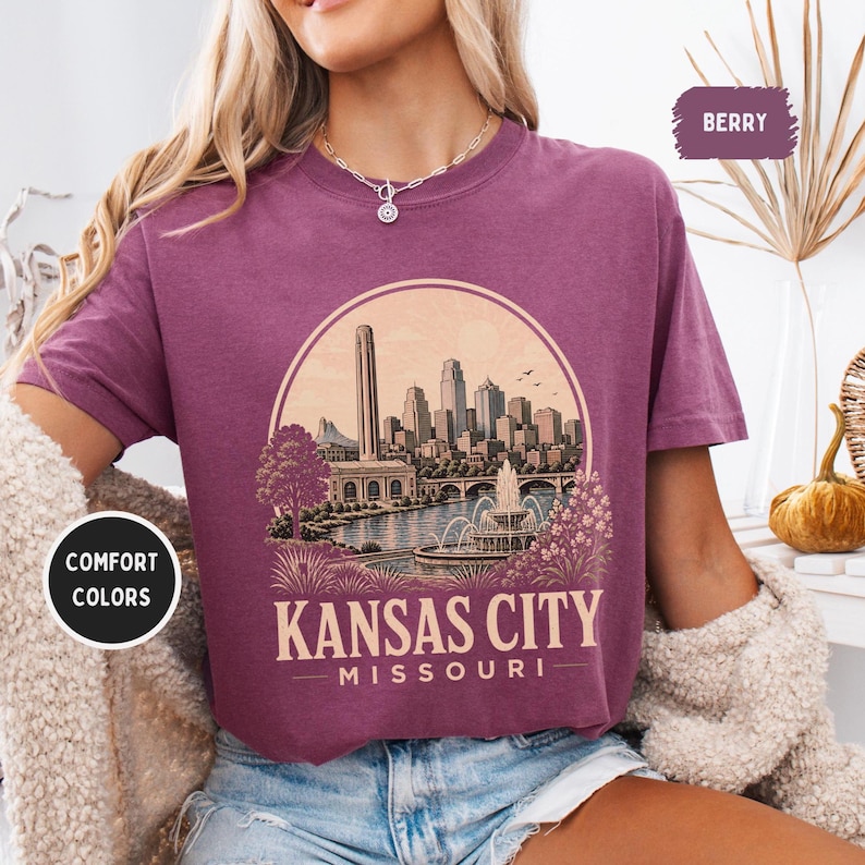 May include: Berry-colored t-shirt featuring a circular graphic of the Kansas City skyline, including a tall tower, buildings, a river, and a fountain. The text "Kansas City Missouri" is below the graphic. The shirt has short sleeves and a crew neck.
