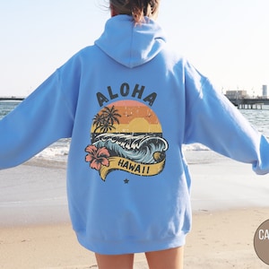 Puede incluir: Una sudadera con capucha azul claro con un estampado gr&aacute;fico vintage de una ola, palmeras y una puesta de sol. El gr&aacute;fico dice "Aloha Hawaii".