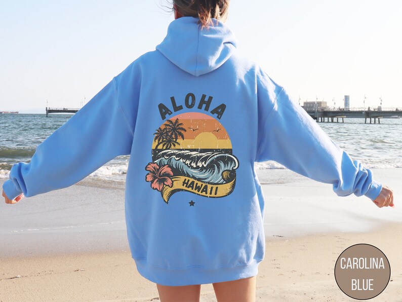 Puede incluir: Una sudadera con capucha azul claro con un estampado gr&aacute;fico vintage de una ola, palmeras y una puesta de sol. El gr&aacute;fico dice "Aloha Hawaii".