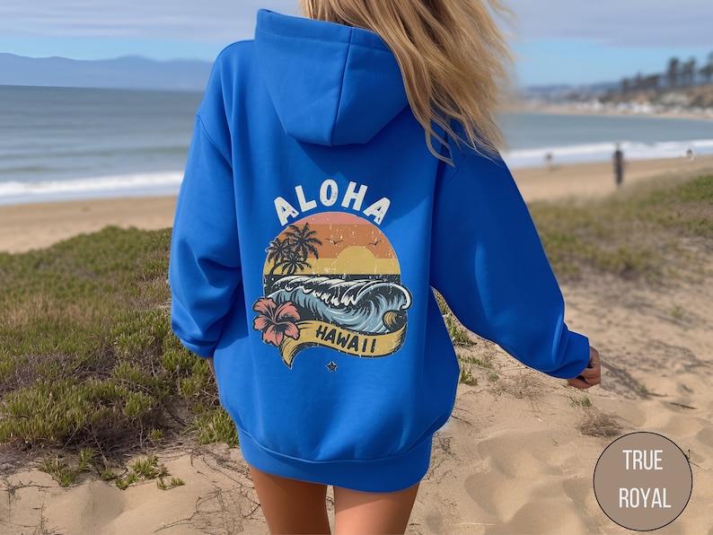 Puede incluir: Una sudadera con capucha azul real con un gr&aacute;fico de estilo vintage en la espalda que dice "Aloha Hawaii" con un dise&ntilde;o de palmera, puesta de sol y ola.