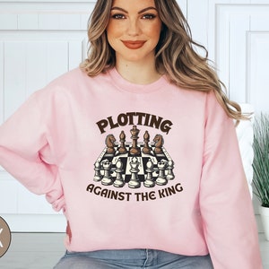 May include: A pink sweatshirt with the text "Plotting Against The King" and a graphic of a chessboard with chess pieces.
