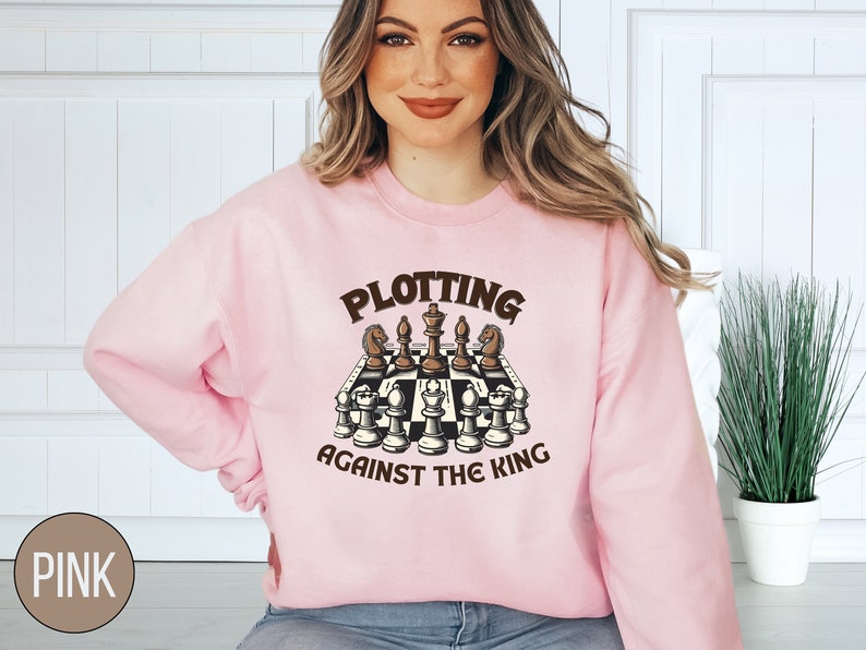 May include: A pink sweatshirt with the text "Plotting Against The King" and a graphic of a chessboard with chess pieces.