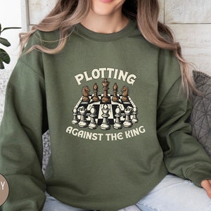 May include: A green sweatshirt with the text "Plotting Against The King" and a graphic of a chessboard with chess pieces.