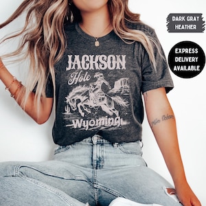 Puede incluir: Camiseta gris oscuro jaspeado con un gráfico desgastado de un vaquero a caballo y el texto "Jackson Hole Wyoming". La camiseta tiene cuello redondo y mangas cortas. También son visibles las palabras "Express Delivery Available".